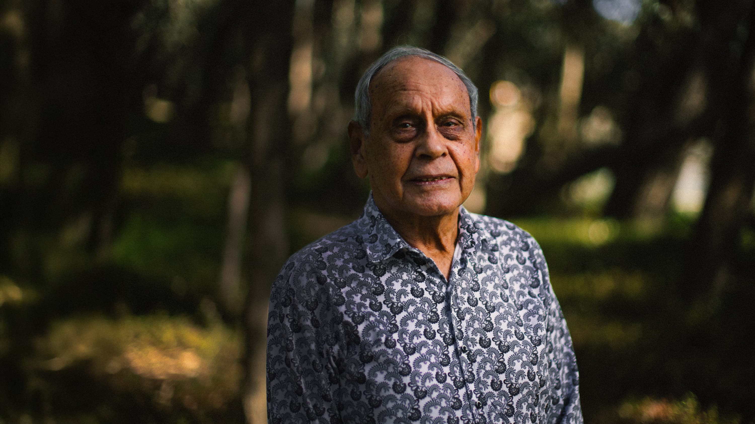 Headshot of Wollithiga Elder Uncle Henry Atkinson wearing a grey checkered shirt, framed by softly blurred green trees in the background.