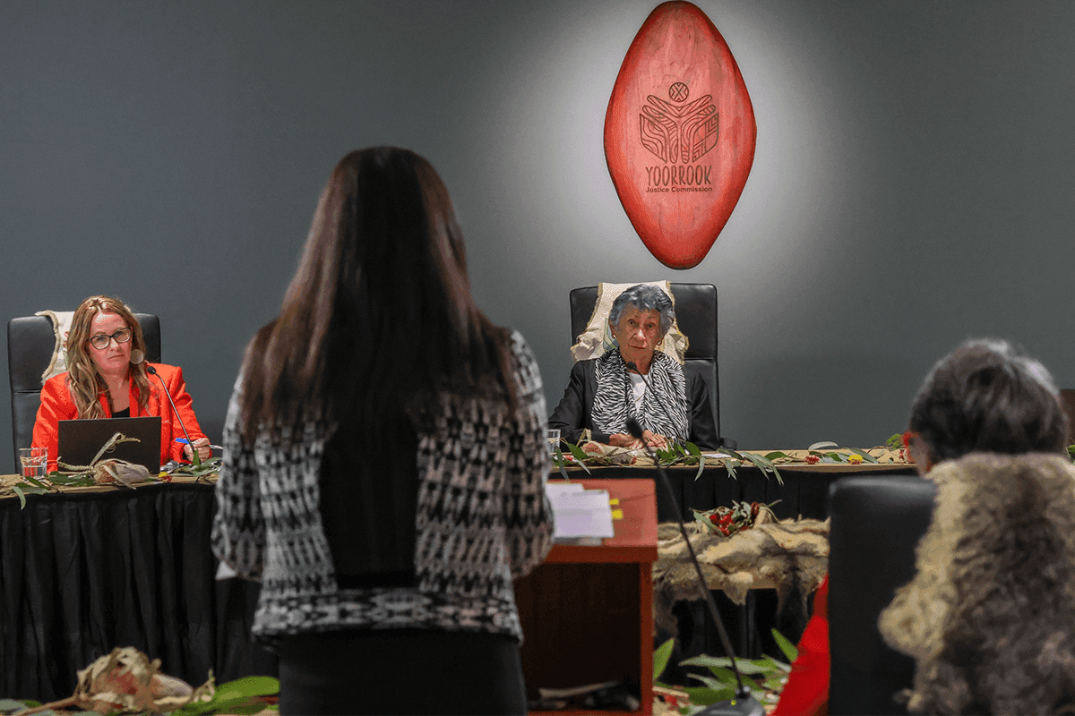 Yoorrook chair Aunty Eleanor Bourke sits center right at a panel table during a formal hearing. A state witness stands in the foreground, addressing the panel. The Yoorrook shield is visible in the background.
